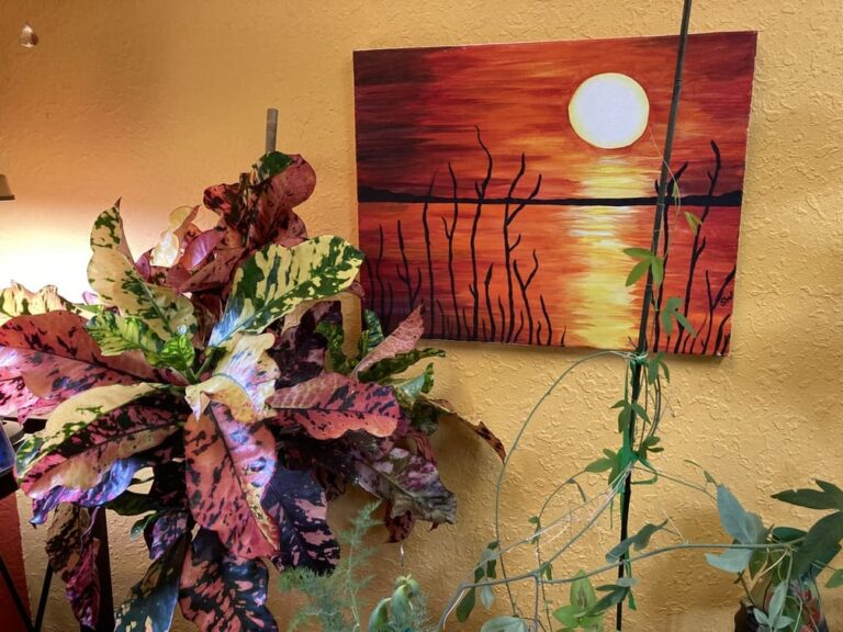 Colorful red, yellow and green croton next to painting with similar color tones against a yellow wall